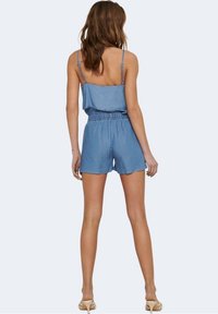 Mono de mezclilla con tirantes de spaghetti, cintura elástica, pantalones cortos acampanados y un detalle de volantes en capas en la parte superior, en un color azul claro.