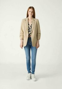 Beiger oversized Blazer über einem gemusterten Oberteil, kombiniert mit blauen Skinny-Jeans und weißen Sneakern. Glattes Material und Knopfdetail an den Jeans.