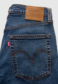 Σκούρο μπλε τζιν παντελόνι με δερμάτινη ετικέτα Levi’s, εμβληματική ραφή πίσω τσέπης και μικρή κόκκινη ετικέτα Levi’s στο πλάι.