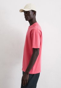 Lichtroze katoenen t-shirt met korte mouwen, ronde hals, zijslits detail, gedragen met donkere broeken en een lichte pet.