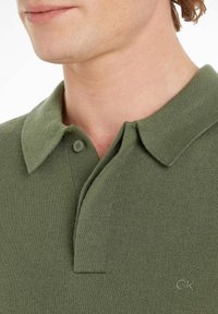 Grünes Poloshirt aus Strickstoff mit geknöpftem Schlitz, flachem Kragen und kleinem Logo auf der unteren rechten Seite.