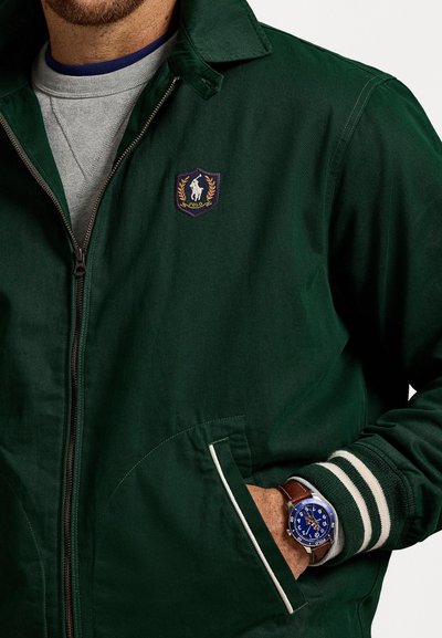 Mand iført grøn zip-up jakke med Polo-logo, grå sweatshirt under, og brunt læderarmbåndsur med blå skive, hånd i lommen.