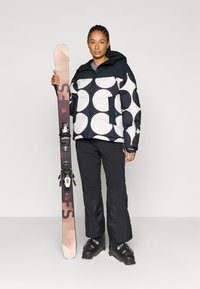 Zwart-witte ski-jas met polkadots, zwarte broek, zwarte skistiefels, met ski's met bindingen in de hand. Gladde textuur met een opgeblazen ontwerp.