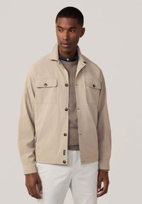 Lichtbeige jacket met een kraag, twee borstzakken, bruine knopen en een ontspannen pasvorm, gedragen over een grijze trui en witte broek.