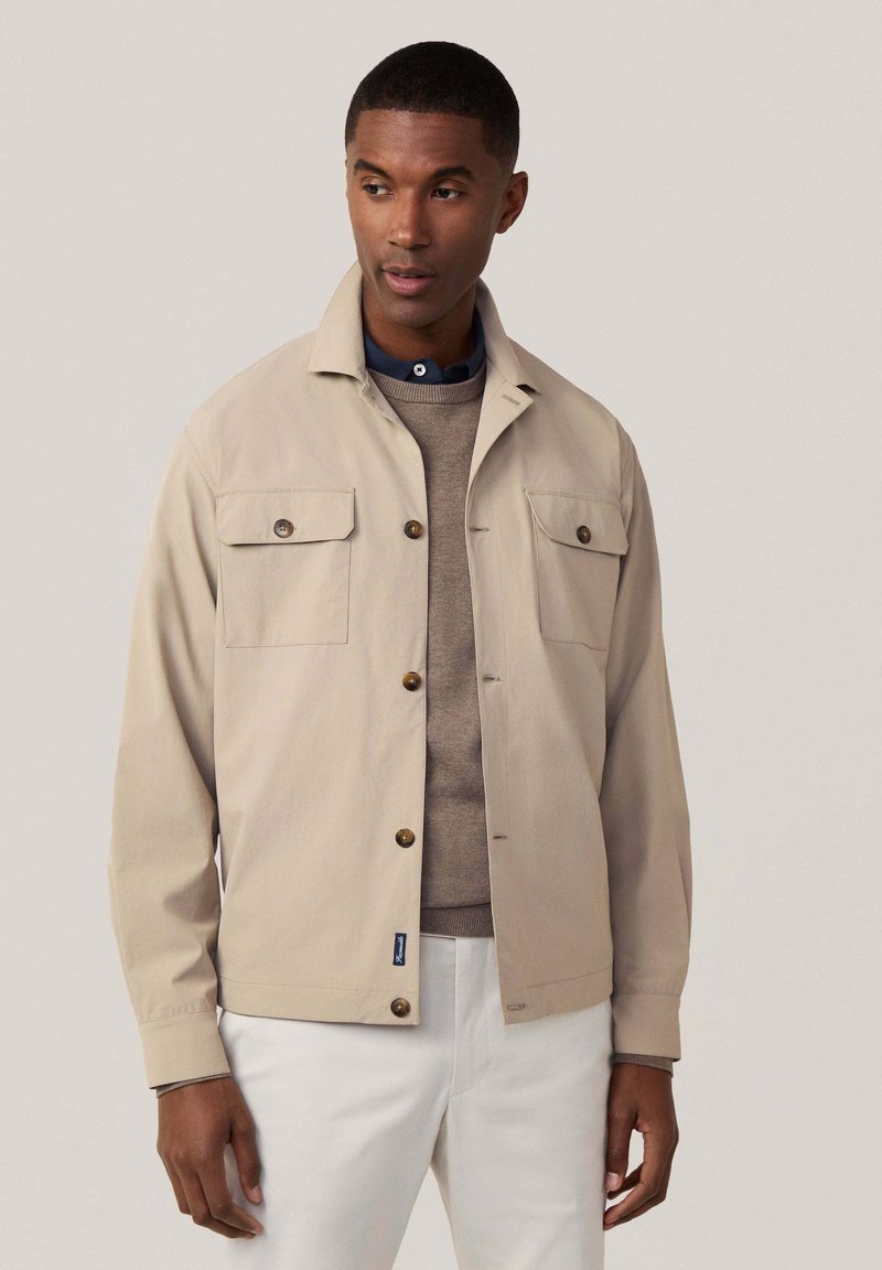 Lichtbeige jacket met een kraag, twee borstzakken, bruine knopen en een ontspannen pasvorm, gedragen over een grijze trui en witte broek.