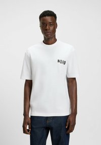 Man wearing a white short-sleeve t-shirt with "Hugo" text on left chest, paired with dark blue jeans, standing against a plain background.