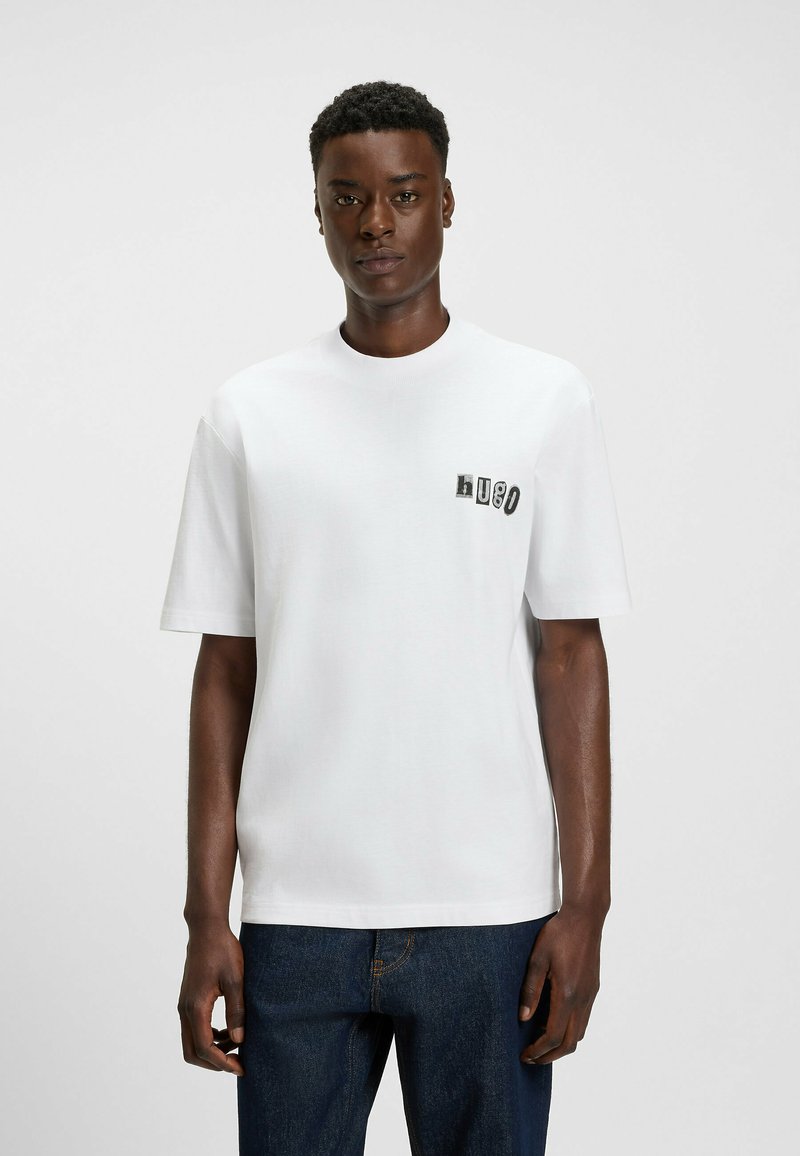 Man wearing a white short-sleeve t-shirt with "Hugo" text on left chest, paired with dark blue jeans, standing against a plain background.