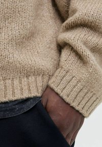 Beiger Strickpullover mit gerippten Bündchen, der eine weiche Textur aufweist, kombiniert mit einer dunklen unteren Schicht. Nahaufnahme des Materials und der Handschrift.