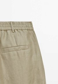 Pantalon beige avec une ceinture élastique et une seule poche passepoilée à l’arrière côté droit.