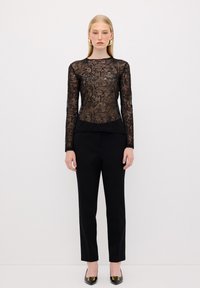Top a maniche lunghe in pizzo nero con motivo floreale, sovrapposto a una base nera solida. Abbinato a pantaloni neri su misura e scarpe nere a punta.