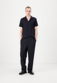 Camicia a maniche corte, con bottoni, di colore blu scuro con una finitura testurizzata, abbinata a pantaloni a gamba larga con righe sottili e scarpe nere.