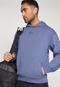 Γκρι-μπλε φούτερ Nike Pro με μπροστινή τσέπη, μακριά μανίκια και κουκούλα με κορδόνι. Το μοντέλο κρατάει μια μαύρη αθλητική τσάντα με λευκό λουρί.