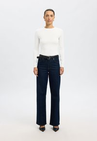 Bela dolgoročna majica združena s temno modrimi kavbojkami z visokim pasom, ki jih krasi črni pas in črne zašiljene čevlje, stoji pred svetlo podlago.