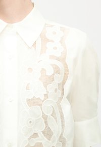 Λευκό πουκάμισο με κουμπιά, με διαφανές μπροστινό πάνελ από δαντέλα με λουλούδια και κοντά μανίκια, εμφανιζόμενο από τον λαιμό έως το μέσο του κορμού.