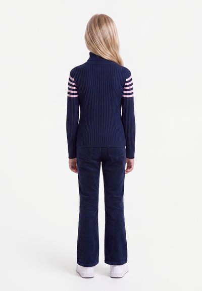Enfant aux longs cheveux blonds portant un pull côtelé bleu marine avec des rayures roses sur les manches, un pantalon bleu marine et des chaussures blanches, debout de dos.