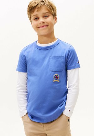 T-shirt in cotone blu con scollatura rotonda, maniche corte e taschino sul petto. Presenta un'emblema ricamato in rosso e oro. Indossato sopra una camicia bianca a maniche lunghe.