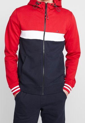 Mann trägt eine Kapuzenjacke mit Reißverschluss, rotem Oberteil, weißem Streifen über der Brust, navyfarbenem Unterteil und passenden navyfarbenen Hosen, die Hände in den Taschen.
