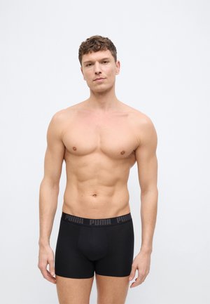 Ing nélküli férfi rövid göndör hajjal, fekete Puma boxeralsót viselve, világos, egyszínű háttér előtt áll.