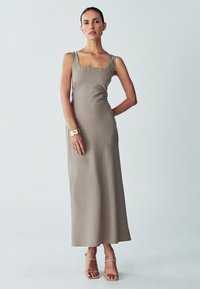 Langes, figurbetontes Kleid in Taupe mit quadratischem Ausschnitt und dünnen Trägern. Aus glattem, mattem Stoff gefertigt. Getragen mit einem goldenen Armband und nude-farbenen Sandalen.