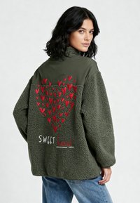 Donna con capelli neri fino alle spalle indossa una giacca oversize in pile verde con motivo a cuori rossi e scritta "SWEET heart" sulla schiena, abbinata a jeans blu.