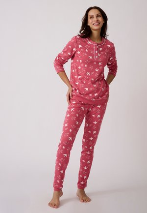 Mujer de pie descalza, vestida con conjunto rosa de manga larga y pantalones con patrones blancos de pájaros, sonriendo y mirando ligeramente hacia arriba.