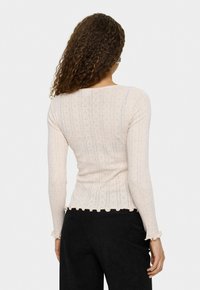 Top roz deschis cu mânecă lungă, cu textură ribbed, tiv ondulat și mici modele în formă de diamant. Materialul pare moale și ajustat.