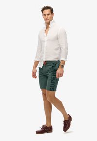 Weißes Leinenhemd mit Kragen, grüne Badehorts mit einem Logo und kastanienbraune Bootsschuhe mit Schnürsenkeln. Oranger Kordelzug an den Shorts.
