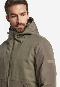 Olivgrön jacka med dragkedja, justerbar huva och strukturerat tyg på övre delen. Logotypmärke på ärmen.