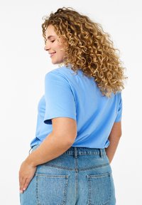 T-shirt blu in cotone a maniche corte con scollatura rotonda, abbinato a jeans in denim a vita alta di un lavaggio chiaro. I capelli ricci incorniciano la vista da dietro.