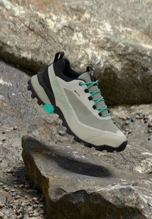 Svetlo sivi planinski čevlji z črnimi in teal akcenti, strukturiran gumijast podplat, ojačan prst in oznaka Gore-Tex za vodoodpornost.