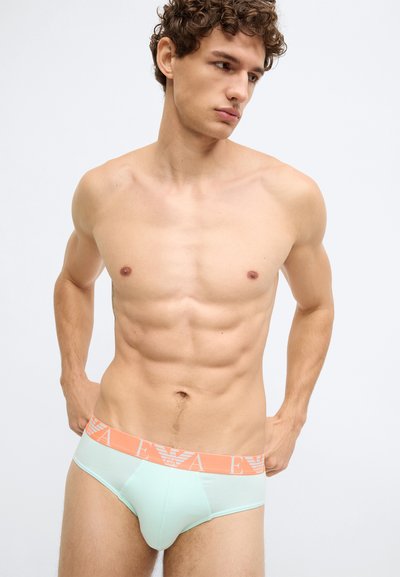 Cueca verde menta com cós laranja apresentando logótipo. O tecido parece liso e ajustado, realçando a forma do corpo.