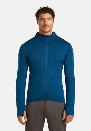 Mies seisoo päällään sininen vetoketjullinen huppari, jossa on peukaloreiät ja rintatasku, yhdistettynä ruskeisiin housuihin valkoista taustaa vasten.