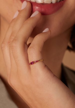 Bague en or avec de petites pierres précieuses roses, dotée d'un bandeau fin et d'un centre en forme d'oval. Main touchant doucement le visage, avec du vernis à ongles neutre.