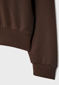 Gros plan d'une manche de vêtement marron avec poignets et ceinture côtelés sur un fond uni et clair.