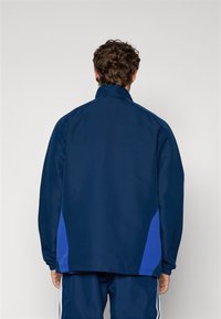 Marineblaue Jacke mit hohem Kragen, aus glattem Stoff gefertigt, mit seitlichen Einsätzen in einem helleren Blau. Lässige Passform mit elastischen Bündchen.