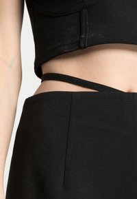 Gros plan sur le ventre d'une personne portant un crop top noir et un pantalon noir taille haute avec une fine sangle croisant la taille.