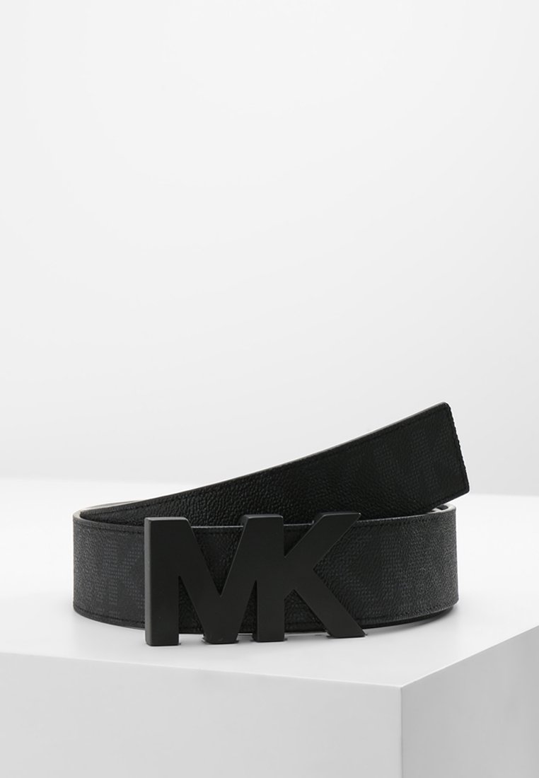 Ceinture en cuir noir avec surface texturée et grande boucle noire présentant les lettres M et K, posée sur une surface blanche.
