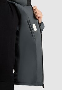 Grijze pufferjack met een gladde, glanzende buitenkant, die de binnenvoering en een binnenzak toont. Bevat een rits en labeldetails.