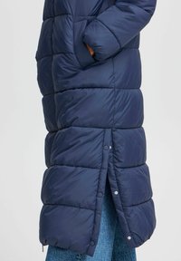 Námornícky prešívaný dlhý kabát s prešívanými časťami, bočnými vreckami, detailmi na patentkách a jemnou textúrou, nosený cez modré denim.