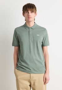 Roheline polo särgid, mis on valmistatud puuvillast, klassikalise kraega, kolme nupukesega ja valge "HUGO" logoga rinnal. Kanda beeži värvi pükste jao.