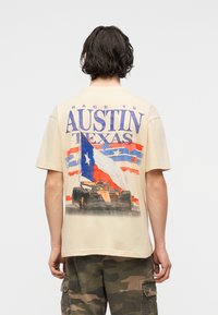 Άτομο με σκούρα μαλλιά που κοιτάζει προς τα πίσω φοράει μπεζ πουκάμισο με το κείμενο "Austin Texas", τη σημαία του Τέξας και γραφικό ενός αγωνιστικού αυτοκινήτου. Φαίνεται να φοράει παντελόνι παραλλαγής.