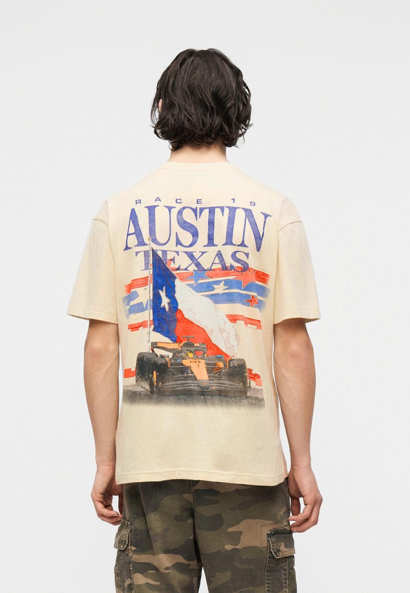 Άτομο με σκούρα μαλλιά που κοιτάζει προς τα πίσω φοράει μπεζ πουκάμισο με το κείμενο "Austin Texas", τη σημαία του Τέξας και γραφικό ενός αγωνιστικού αυτοκινήτου. Φαίνεται να φοράει παντελόνι παραλλαγής.