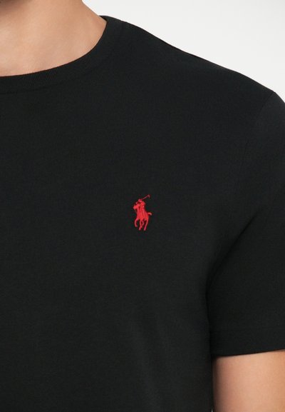 Juodas medvilnės marškinėlis su apvaliu iškirpimu, kuriame kairiajame krūtinės šone yra mažas raudonas išsiuvinėtas logotipas su arklio ir raitelio atvaizdu.