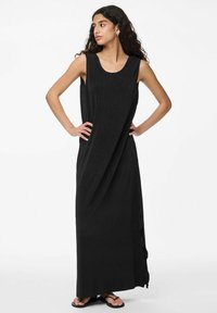 Juodas maksi suknelė su apvaliu iškirpimu, be rankovių ir tekstūruotu audiniu. Pasižymi šoninėmis iškirpėmis, užtikrinančiomis patogumą judant. Avimos plokščios šlepetės.