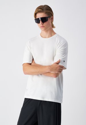Weißes T-Shirt mit kurzen Ärmeln und rundem Halsausschnitt sowie dezentem Logo-Detail am Ärmel, kombiniert mit schwarzen Sportshorts und dunklen Sonnenbrillen.