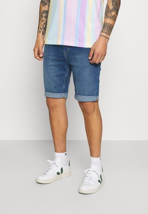 Jeans-Shorts mit umgeschlagenen Säumen, in einem mittleren Blauton gewaschen. Getragen mit einem mehrfarbigen gestreiften T-Shirt und weißen Sneakers.