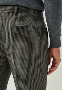 Brown and black houndstooth patterned trousers with a back pocket and button detail, made from a textured fabric.
