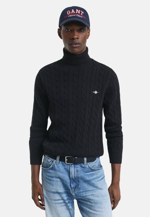 Black cable-knit sweater with high collar, paired with light blue jeans and a navy cap featuring "GANT" in red lettering.
