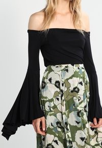 Kvinna som bär en av-axlar svart topp med långa utsvängda ärmar och en högmidjad kjol med grön och vit blommönster.