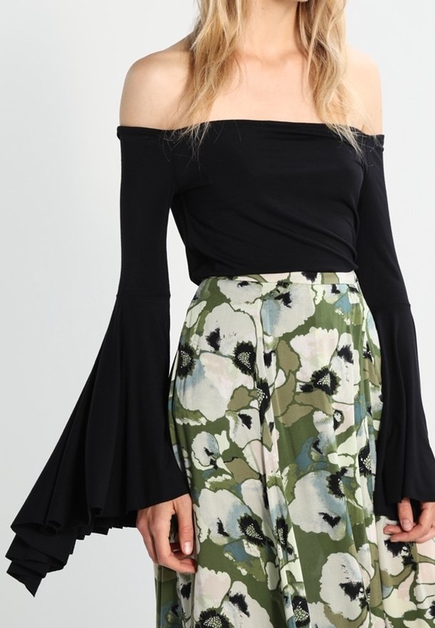 Kvinna som bär en av-axlar svart topp med långa utsvängda ärmar och en högmidjad kjol med grön och vit blommönster.